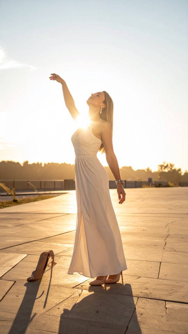 A woman turning into low evening sun, one arm raised, cream dress lifting